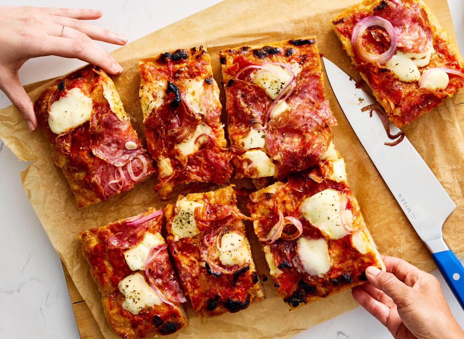 A top-down shot shows a close-up of a person's hands picking up slices of pizza from a sheet of parchment paper. The person on the left is holding a rectangular slice of pizza topped with red sauce, pepperoni, thin red onion slices, and fresh mozzarella. The person on the right is picking up a slice from the bottom right corner of the image. An ACUTO440 8 inch chef's knife with a blue handle, labeled "MISEN," lies on the parchment paper, with a few stray pieces of red onion near its blade.