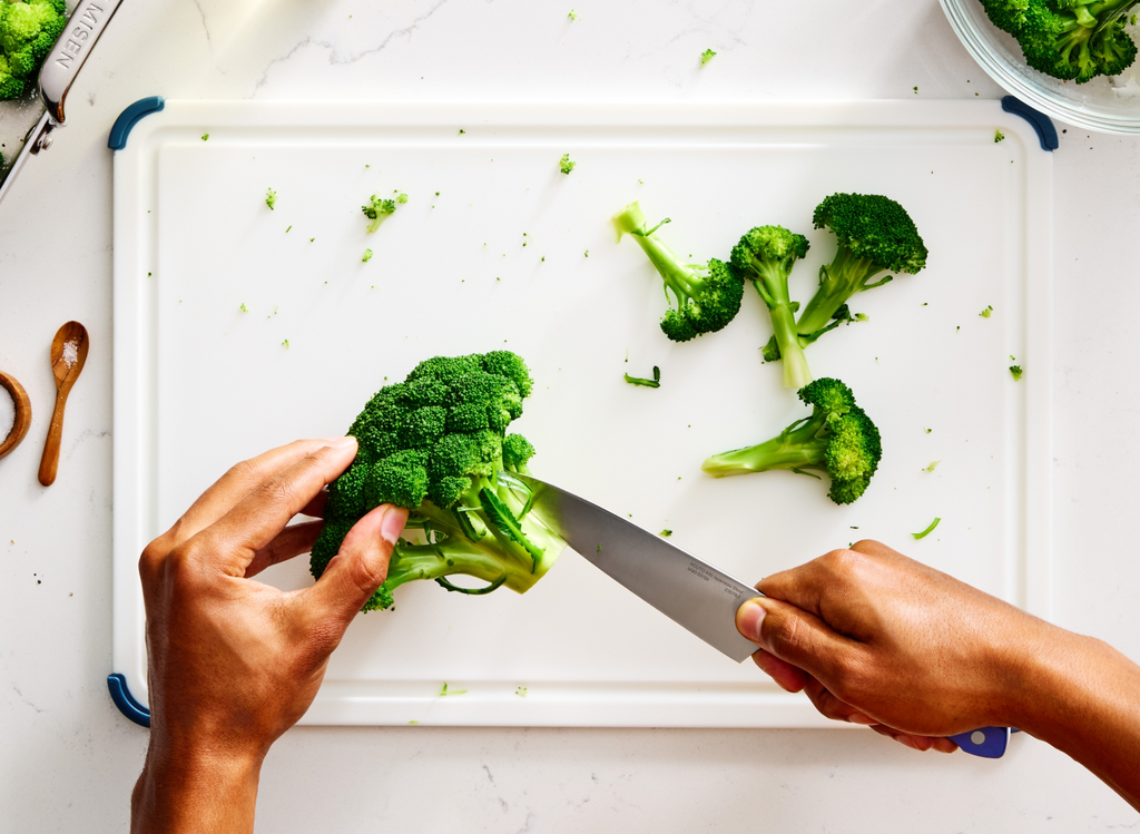 A top-down view shows a person's hands slicing a head of broccoli on a white cutting board. The 5.5" ACUTO440 utility knife, with "MISEN" engraved on the blade, is positioned to cut the florets from the stem. Several broccoli florets are scattered across the right side of the cutting board. In the upper left, a Misen roasting pan holds more broccoli. To the left of the cutting board, a small wooden spoon with a pinch of salt is visible. In the upper right corner, a clear bowl contains more broccoli florets.