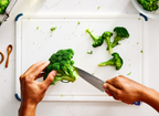 A top-down view shows a person's hands slicing a head of broccoli on a white cutting board. The 5.5" ACUTO440 utility knife, with "MISEN" engraved on the blade, is positioned to cut the florets from the stem. Several broccoli florets are scattered across the right side of the cutting board. In the upper left, a Misen roasting pan holds more broccoli. To the left of the cutting board, a small wooden spoon with a pinch of salt is visible. In the upper right corner, a clear bowl contains more broccoli florets.