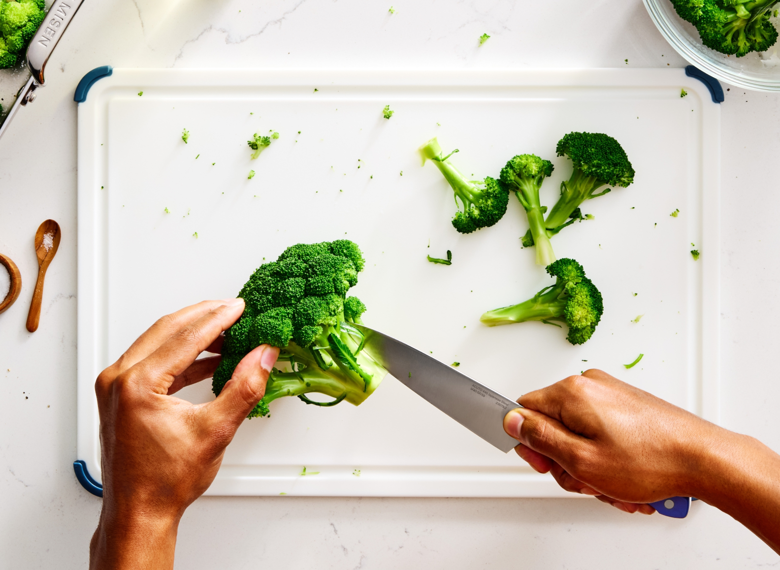 A top-down view shows a person's hands slicing a head of broccoli on a white cutting board. The 5.5" ACUTO440 utility knife, with "MISEN" engraved on the blade, is positioned to cut the florets from the stem. Several broccoli florets are scattered across the right side of the cutting board. In the upper left, a Misen roasting pan holds more broccoli. To the left of the cutting board, a small wooden spoon with a pinch of salt is visible. In the upper right corner, a clear bowl contains more broccoli florets.