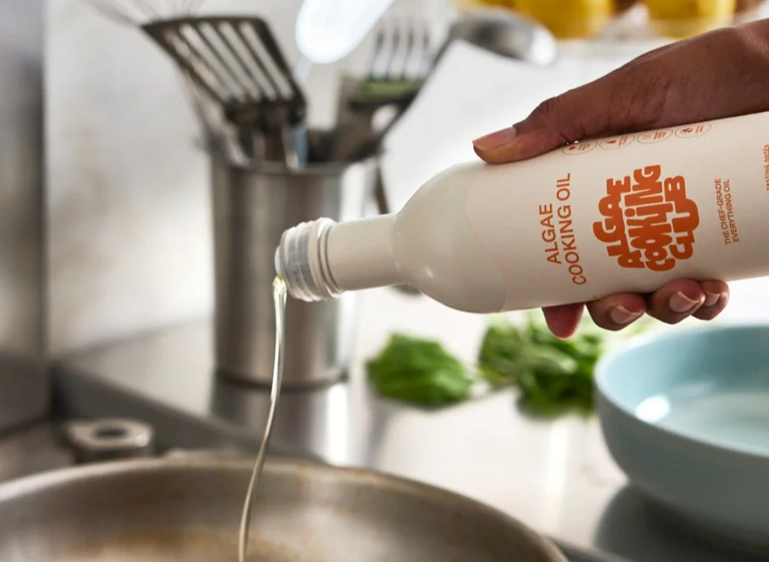 A hand is shown pouring Algae Cooking Oil from a white bottle into a frying pan on a stovetop. The oil forms a steady stream. The bottle's label displays "ALGAE COOKING OIL" and "ALGAE COOKING CLUB" logo in orange, with "THE CHEF-GRADE EVERYTHING OIL" in smaller text above the logo. The background is a blurred kitchen setting with stainless steel tools and a light blue bowl visible.