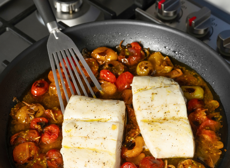 Two seasoned fillets of white fish and cherry tomatoes are cooking in a Misen Carbon Nonstick™ Frying Pan on a stovetop, with a Misen Stainless Steel  Fish Spatula lifting the fish from the pan.
