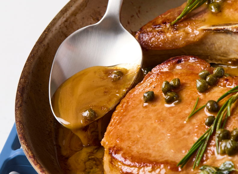 A Misen plating spoon is used to baste two thick, cooked pork chops in a pan. The pork chops are glistening with a rich, golden sauce and garnished with capers and sprigs of fresh rosemary and what appears to be thyme. The pan has a light-colored, rustic-looking exterior and is sitting on a blue Misen silicone roasting rack.