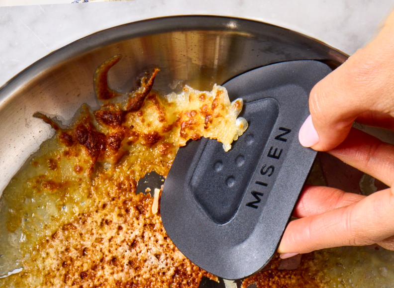 A person's hand uses a black Misen Pan Scraper to remove burnt food from the bottom of a stainless steel saucier. The saucier contains a large amount of burnt, golden-brown cheese.