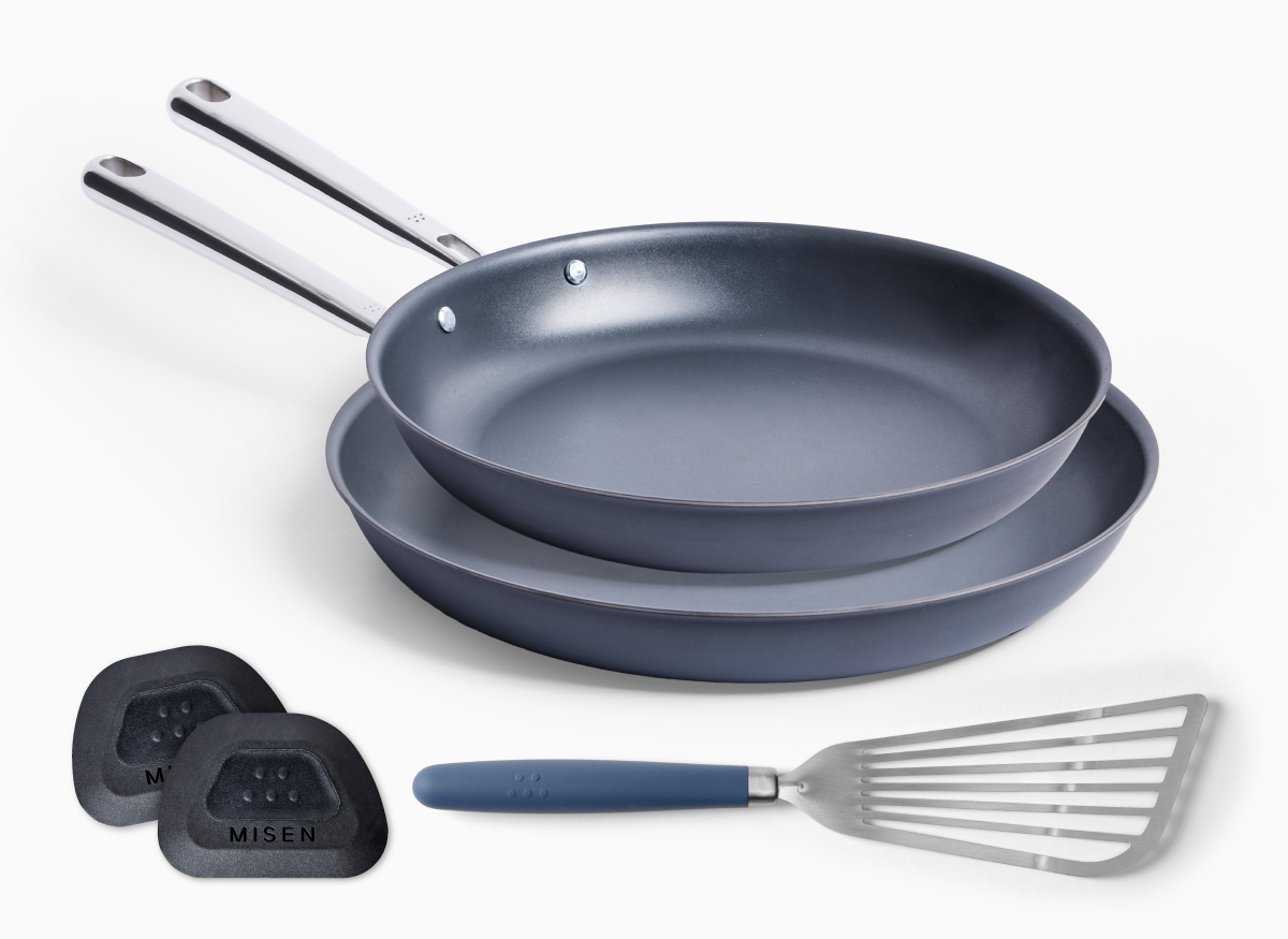 The image depicts a set of Misen nonstick cooking essentials. It includes two Carbon Nonstick™ Frying Pans—one stacked on top of the other—with metallic handles. Below the pans, there is a stainless steel fish spatula with a blue handle, and two pan scrapers, which has "MISEN" text on it. The items are set against a white background.
