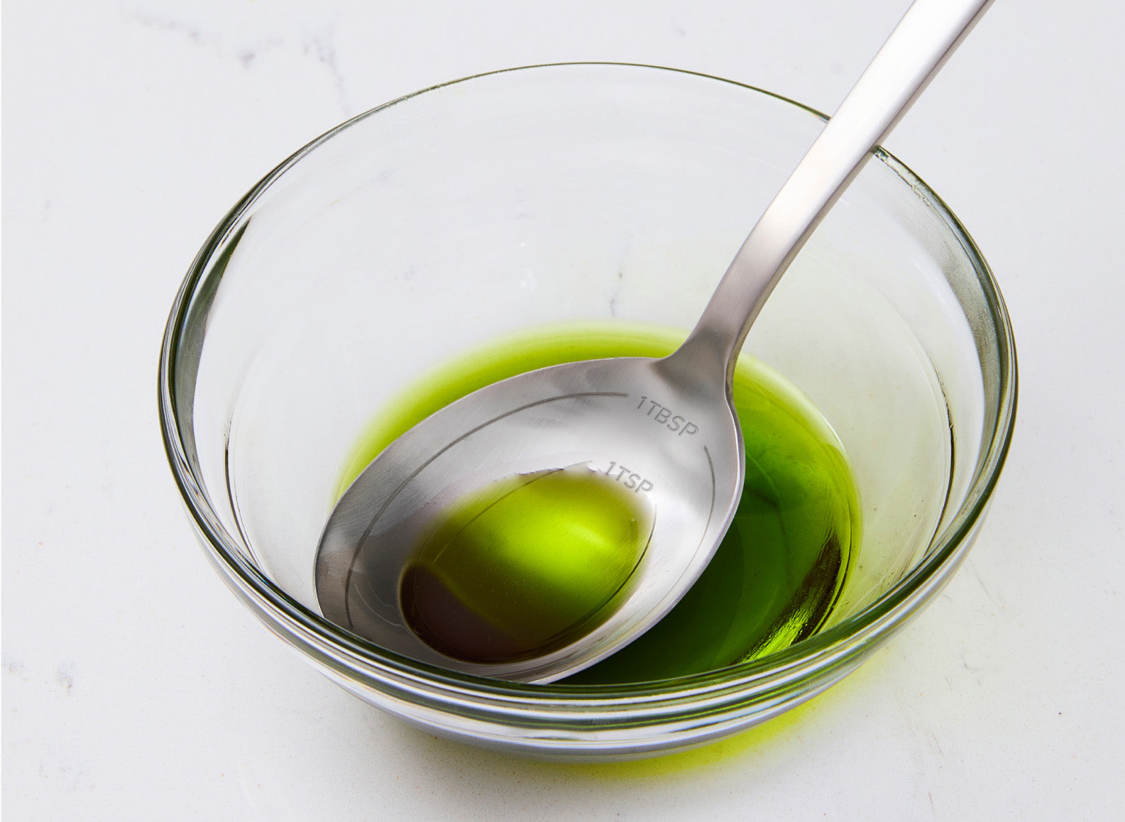 A close-up of a clear glass bowl filled with green extra virgin olive oil. A Misen Measuring Spoon is resting in the bowl, partially submerged in the oil. The background is a white marble countertop.