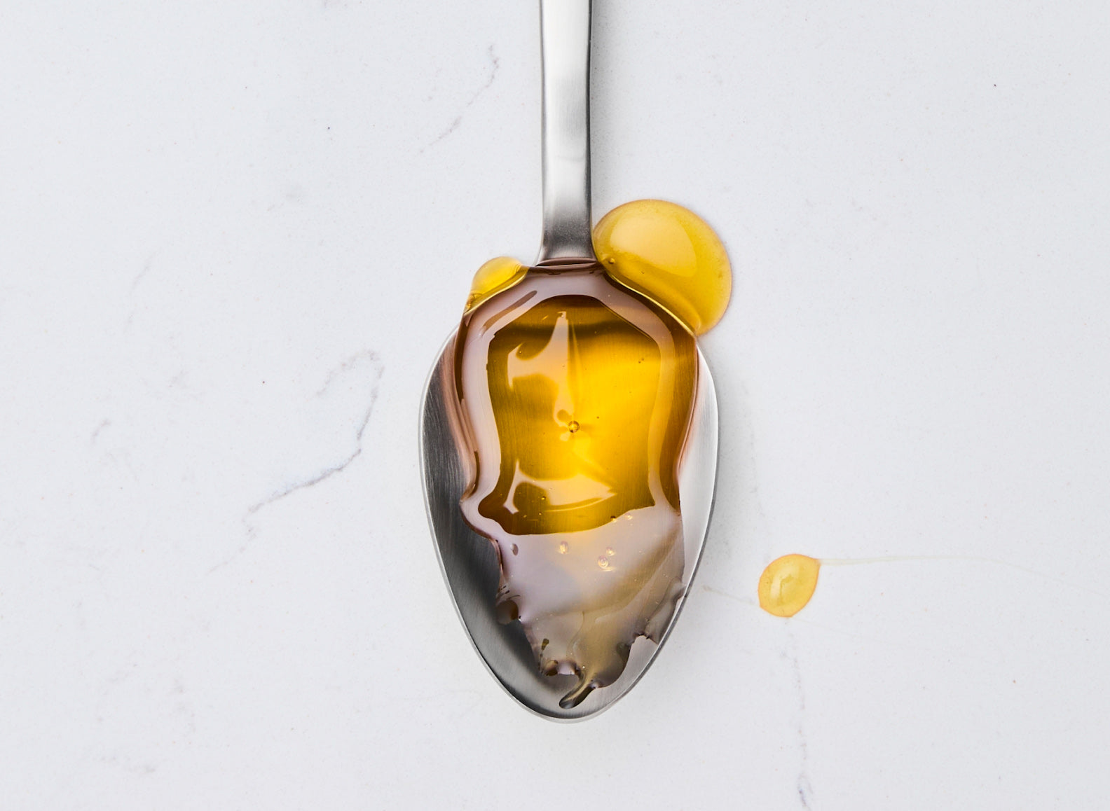 A close-up view shows a Misen stainless steel plating spoon, positioned vertically on a white marble countertop. The spoon's shallow, oval-shaped bowl is filled with golden honey, which also drips slightly onto the marble to the right of the spoon. The honey creates reflections on the polished surface of the spoon.
