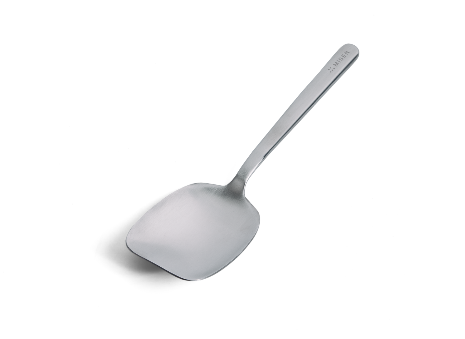 A Misen stainless steel scraper-spoon is positioned diagonally against a white background, with its handle extending towards the upper right. The spoon has a wide, flat, and somewhat rectangular head with gently curved edges, designed for scraping. The handle is slender and widens slightly near the head. The word "MISEN" is subtly debossed on the upper right side of the handle, near its end.