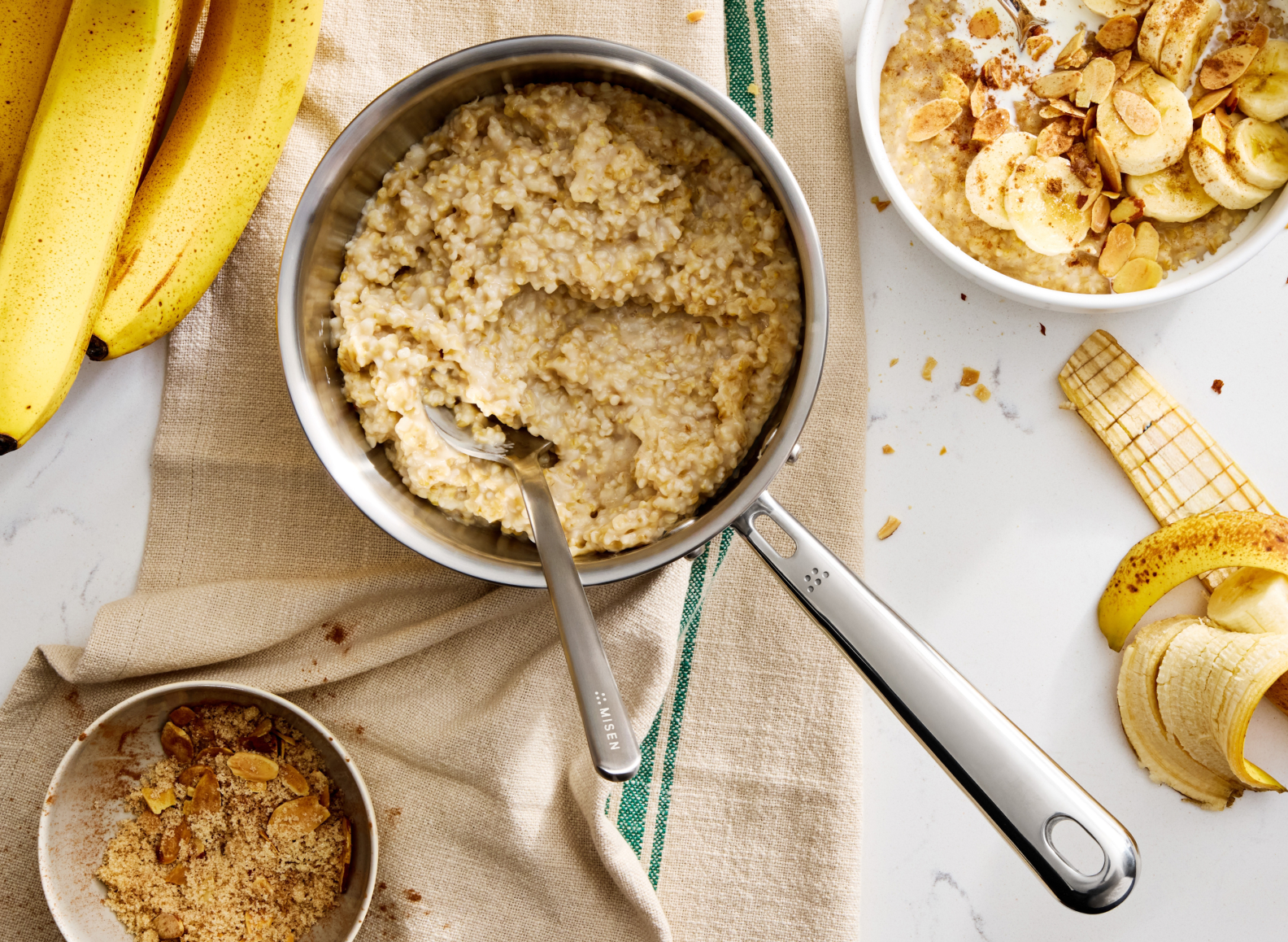 An overhead studio shot shows a stainless steel Misen Saucier filled with cooked quinoa porridge. A plating chef spoon, with "MISEN" engraved on the handle, rests in the saucier. The saucier sits on a green-striped kitchen towel, which is on a white marble countertop. To the left is a bunch of ripe yellow bananas. In the upper right corner is a white bowl of the same porridge, garnished with sliced bananas and almonds. In the bottom left is a small brown bowl of toasted almonds and brown sugar.