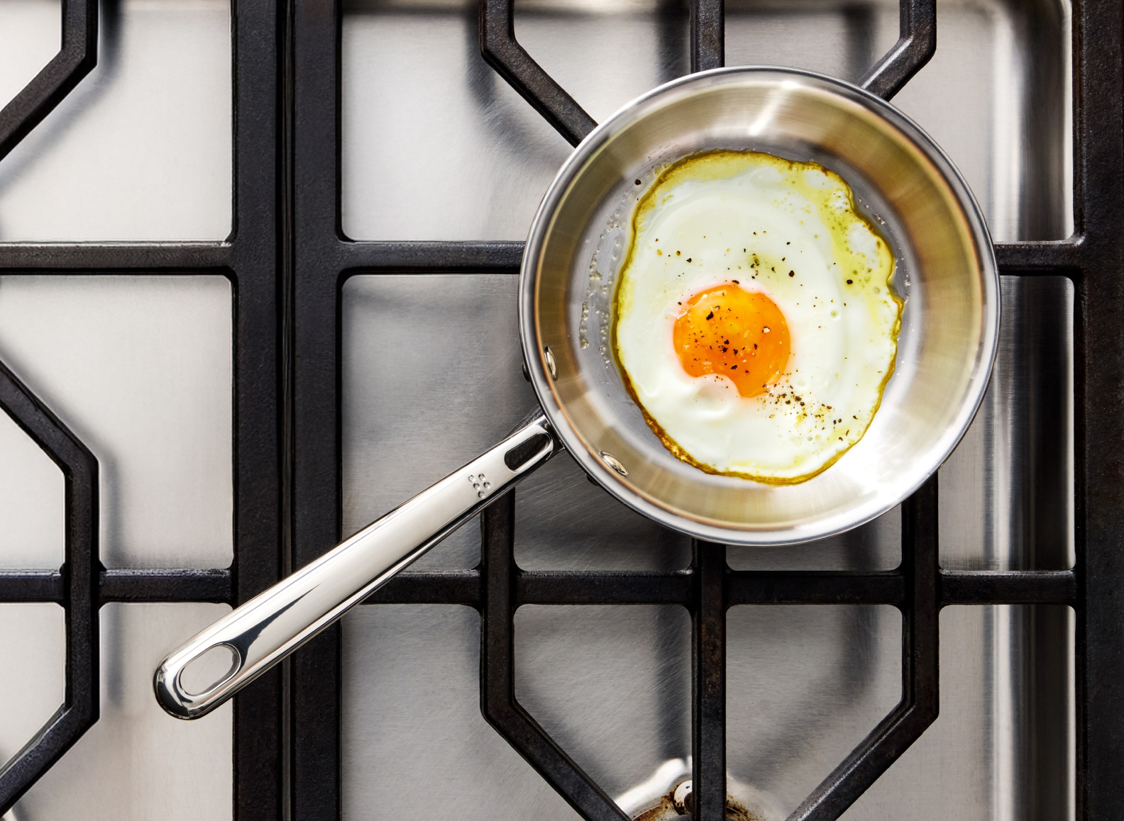 A close-up view of a Misen stainless steel frying pan with a single, perfectly fried egg (sunny-side up with pepper) resting on a black gas stovetop grate.