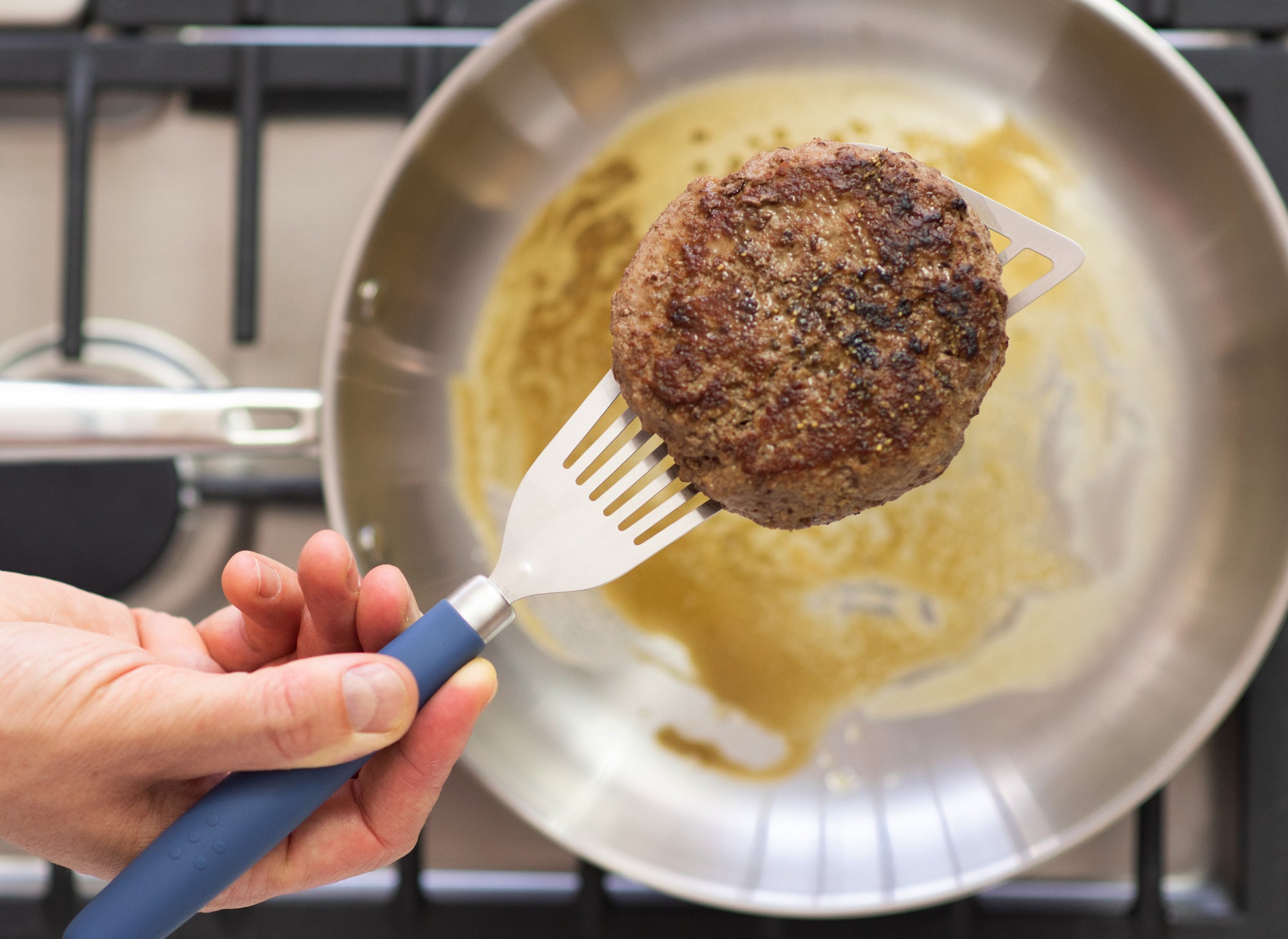 A Misen Metal Fish Spatula with blue silicone handle lifting a cooked hamburger patty above a stainless steel skillet.