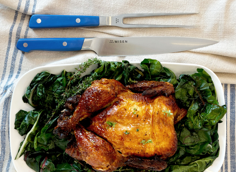A golden brown roasted chicken seasoned with herbs, sitting on a bed of greens in a white rectangular dish. Above the dish are a carving knife and fork, both with blue handles, and the word "MISEN" is visible on the knife.