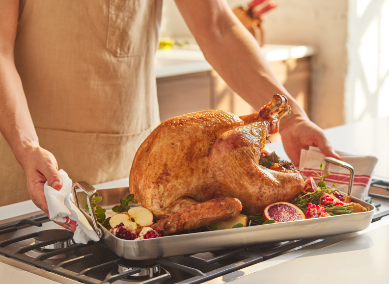 A person wearing a tan apron is holding a large, golden-brown roasted turkey in a Misen Roasting Pan. The turkey is garnished with herbs, sliced apples, pomegranates, and blood oranges. The pan is resting on a stovetop with grates.