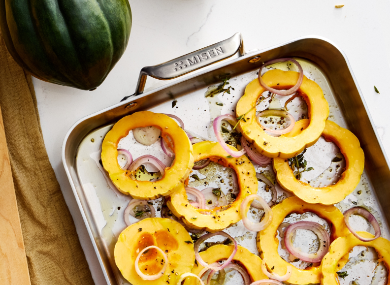 An overhead shot of a Misen Roasting Pan filled with ring-shaped slices of acorn squash and red onion, seasoned with herbs and oil. The word "MISEN" is visible on the handle of the pan. A dark green squash is partially visible in the upper left corner.