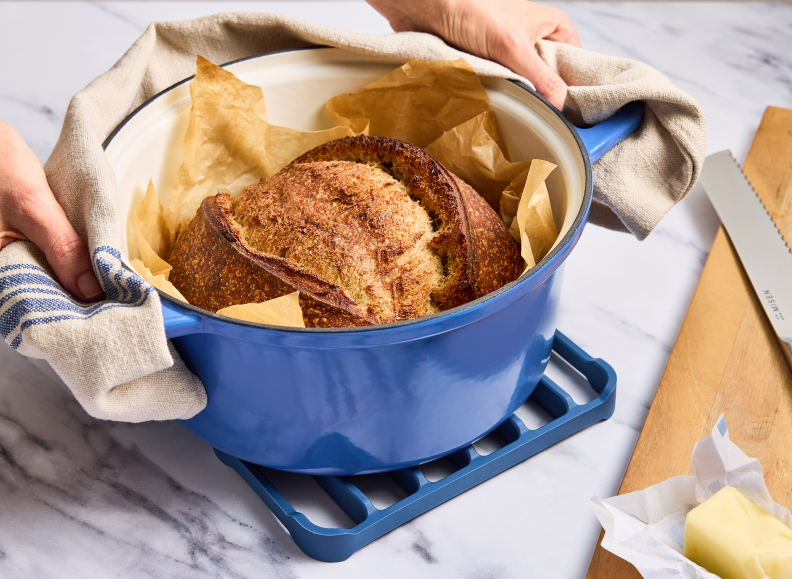 A pair of hands wrapped in a blue Misen Kitchen Towel are holding a blue Misen Dutch Oven containing a loaf of bread baked on parchment paper. The Dutch Oven is sitting on a blue Misen Silicone Roasting Rack on a white marble surface. To the right, a Misen Serrated Knife and a stick of butter are visible on a wooden cutting board. The word "MISEN" is visible on the knife.
