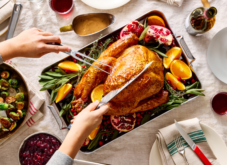 A close-up, top-down view shows a pair of hands carving a golden-brown roasted turkey in a Misen stainless steel 18 inch Roasting Pan. The turkey is garnished with orange slices, pomegranate seeds, and fresh sage sprigs. To the left, a Misen Stainless Steel Frying Pan of roasted Brussels sprouts is visible, and to the lower left, a Misen Saucier of cranberry sauce. A gravy boat, an opened bottle of wine, and glasses of wine are also on the table, which is covered with a light-colored linen tablecloth.