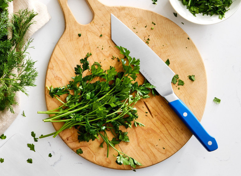 A bundle of parsley sits on a light wooden cutting board next to a blue-handled Misen 6 inch Bunka Knife. A small white bowl with chopped herbs is visible in the upper right corner, and a beige kitchen towel with more herbs is in the upper left.
