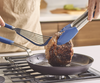 Flipping a pork chop with large blue Misen Silicone Tongs and a blue silicone Misen Fish Spatula in a Misen Pre-Seasoned Carbon Steel Pan.