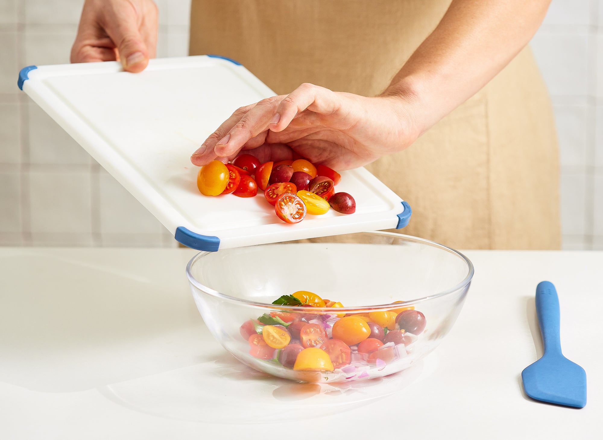 A chef scrapes halved cherry tomatoes off a small Misen Plastic Cutting Board and into a glass bowl.