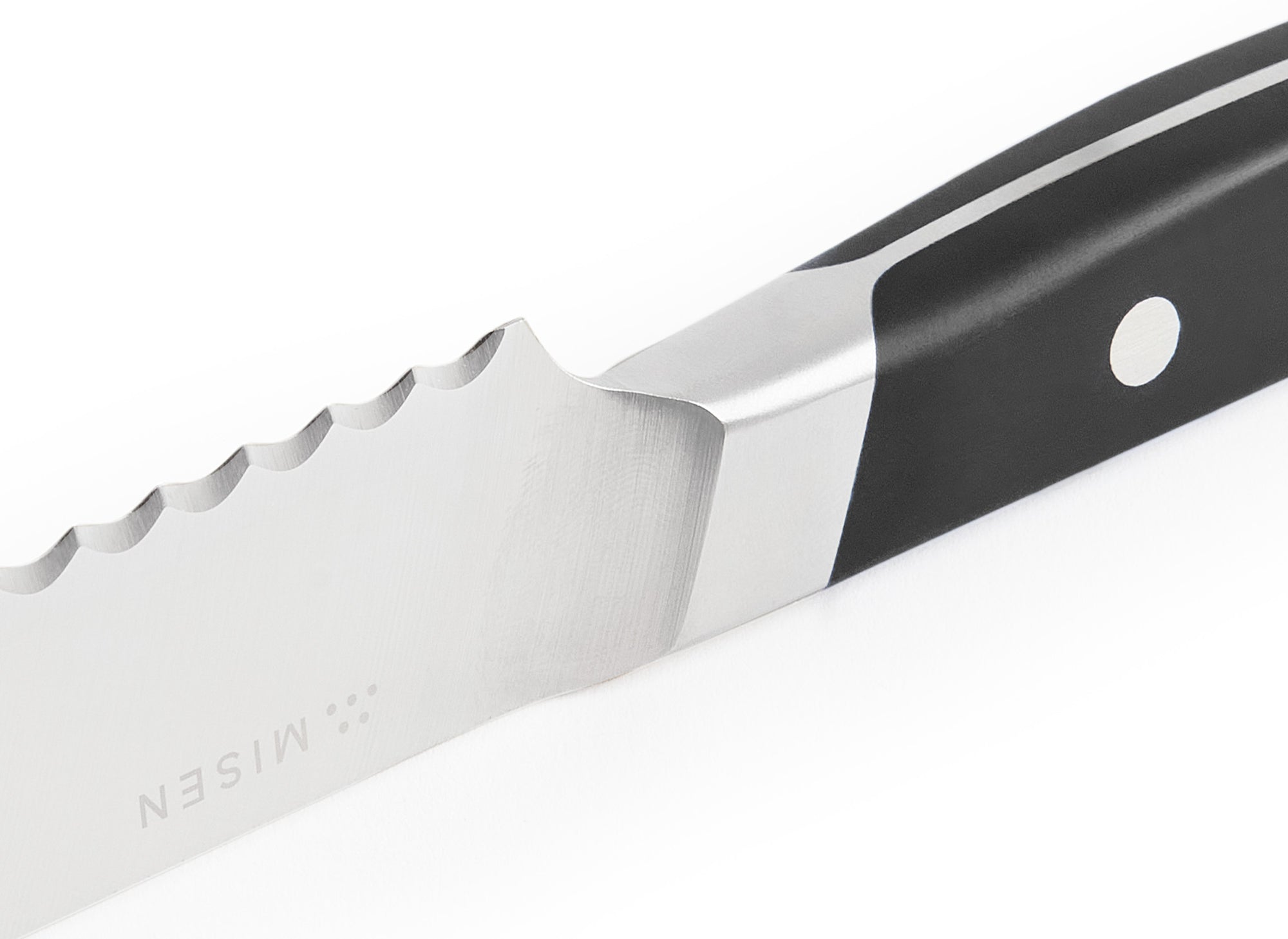 A close-up shot of the black Misen Short Serrated Knife on a white background, with a clear view of the blade serrations and sloped bolster. The blade is facing up and toward the viewer.
