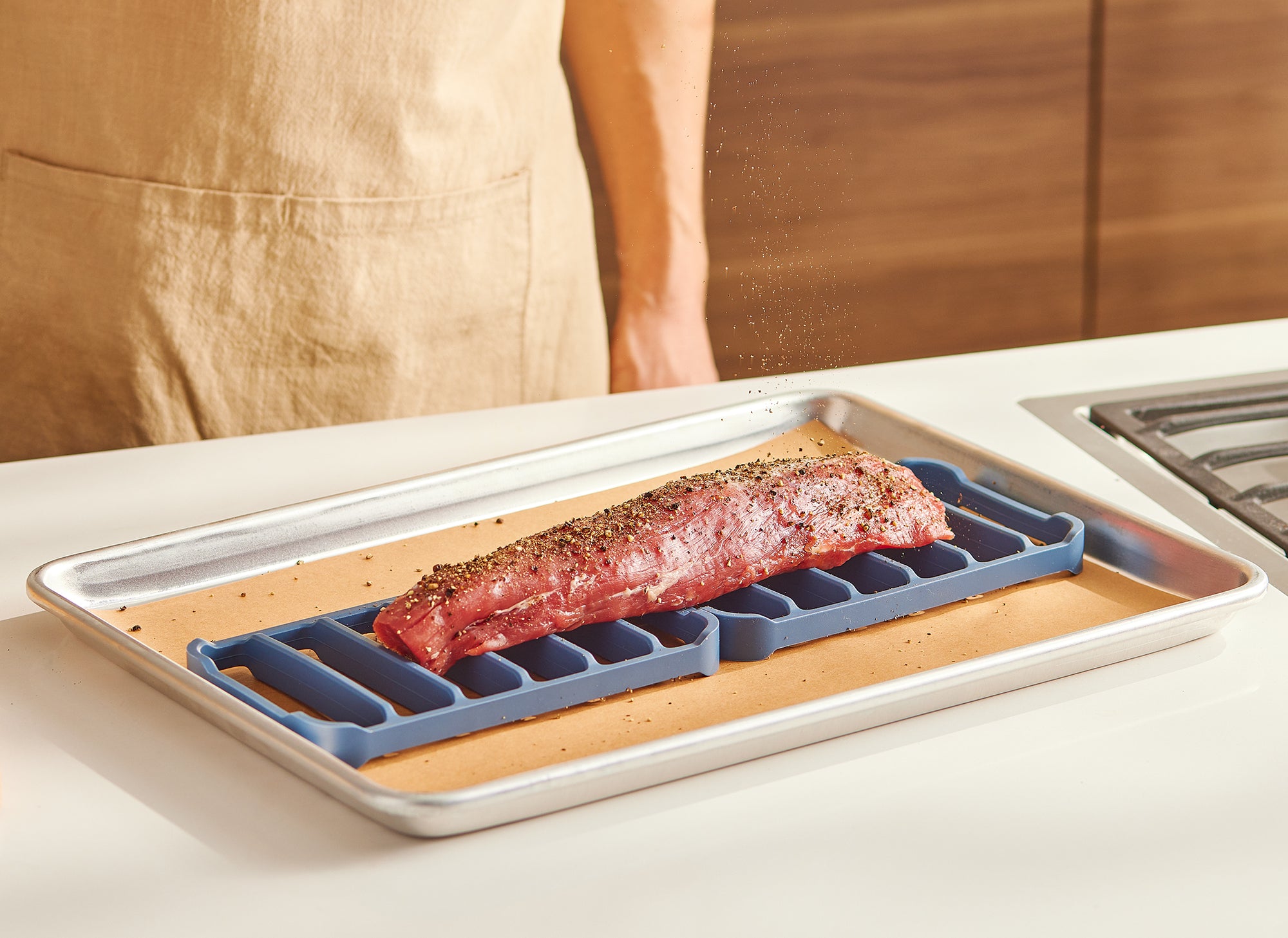 A chef seasons a pork loin, which is sitting atop two blue Misen Silicone Roasting Racks in a parchment paper-lined baking pan.