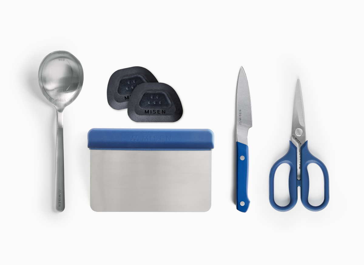 A top-down view shows a collection of cooking tools, including a Misen Measuring Chef Spoon, two Misen Pan Scrapers, a rectangular Misen Bench Scraper with a blue handle, a blue-handled Misen 3.5 inch Paring Knife 2.0, and a pair of blue-handled Misen Kitchen Shears. All items are branded with the name "MISEN." The measuring chef spoon is engraved with "1TBSP SML," and the knife's ACUTO440 steel blade and small shears also have brand markings.
