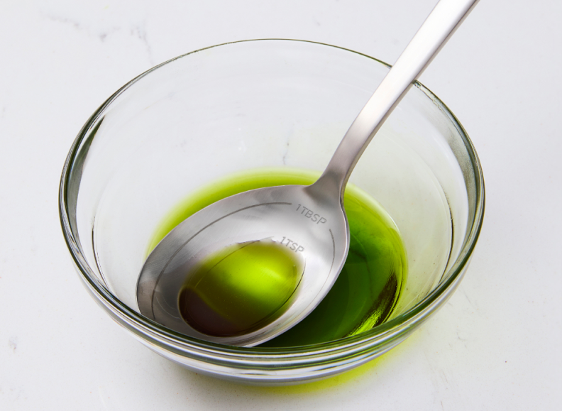 A stainless steel Misen Measuring Chef Spoon labeled "1 TBSP" and "1 TSP" sits inside a clear glass bowl containing green olive oil.