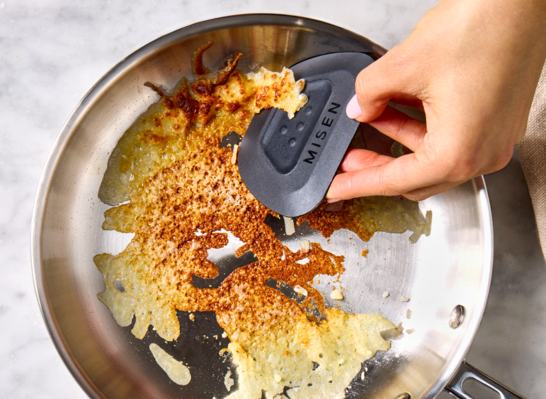 A person's hand uses a black Misen Pan Scraper to remove burnt food from the bottom of a stainless steel saucier. The saucier contains a large amount of burnt, golden-brown cheese.