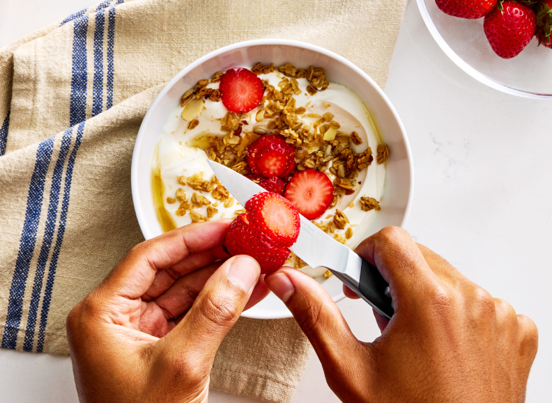 A person's hands are shown from above, slicing a strawberry with a 3.5 inch black Misen Paring Knife 2.0 over a white bowl filled with yogurt, granola, sliced almonds, and strawberries. A blue striped Misen Kitchen Towel is to the left of the bowl, and a clear bowl with whole strawberries is visible in the upper right corner.