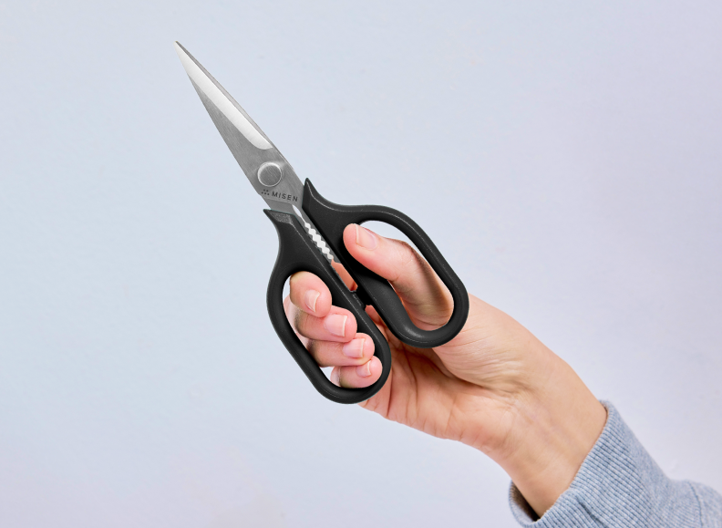 A person's hand holds a black-handled pair of small Misen Kitchen Shears, with the blades pointing upwards and slightly to the left. The Misen logo is visible near the pivot screw of the shears.