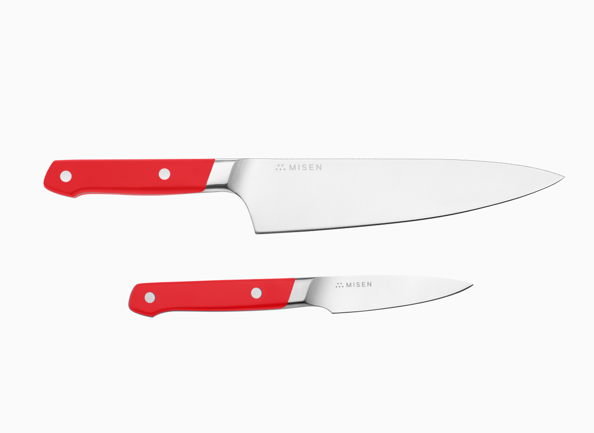 A set of two kitchen knives with red handles and silver blades, one Chef's, one Paring, laid on a white surface.
