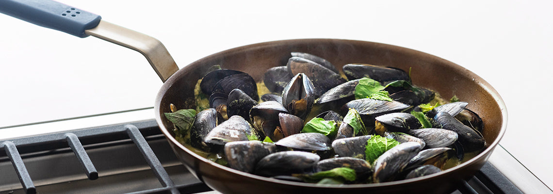 A Misen Carbon Steel Pan on a stovetop filled with steamed and seasoned mussels with herbs.