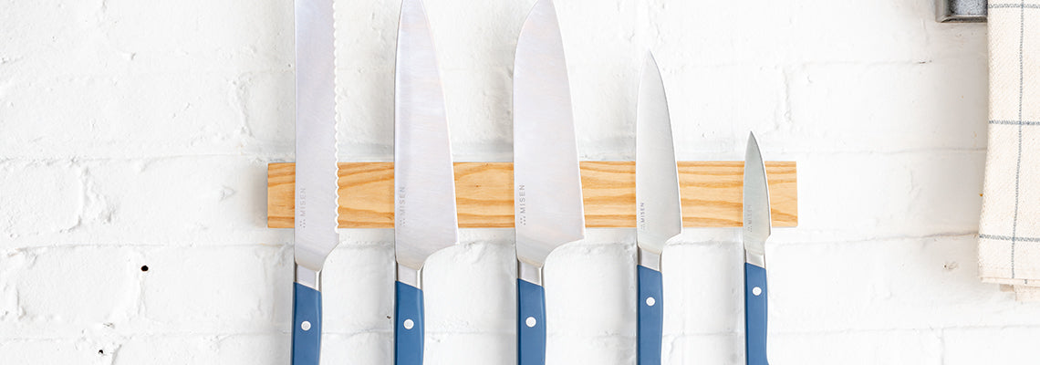 A straight-on view of the Misen 5-Piece Essentials Knife Set mounted on the Misen Magnetic Knife Strip on a wall. From left to right: the blue Serrated Knife, the blue Chef’s Knife, the blue Santoku Knife, the blue Utility Knife, the blue Paring Knife.