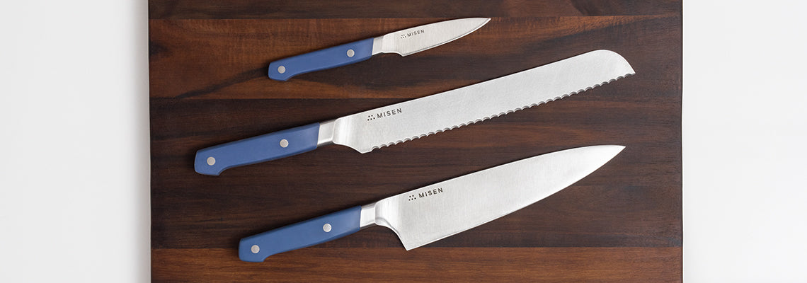 A bird’s eye view of the Misen 3-Piece Essentials Knife Set on a wood Misen cutting board. From top: the blue Paring Knife, the blue Serrated Knife, and the blue Chef’s Knife.