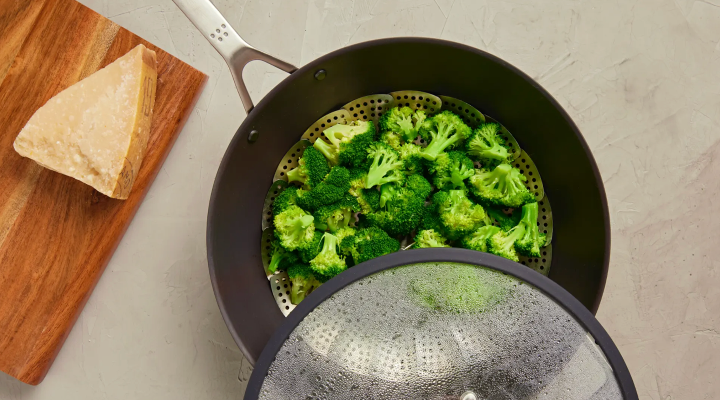 Fresh broccoli florets steaming in a basket inside a dark nonstick pan with a glass lid and parmesan cheese on a wooden cutting board