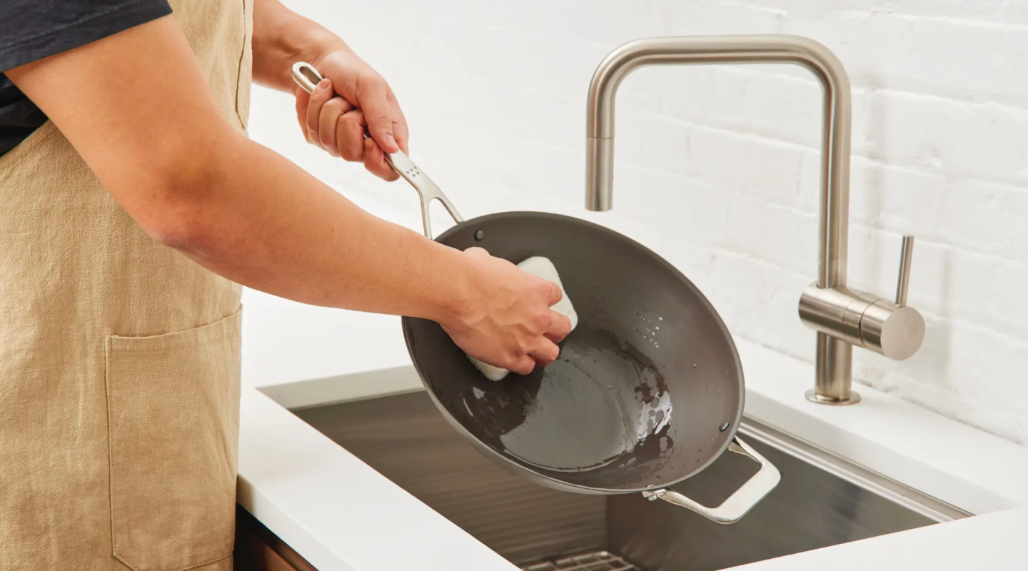Person in apron scrubbing a dark nonstick wok pan with a sponge at kitchen sink