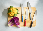 Wood cutting board with five Misen chef knives with colored handles, yellow cauliflower, and halved purple cabbage.