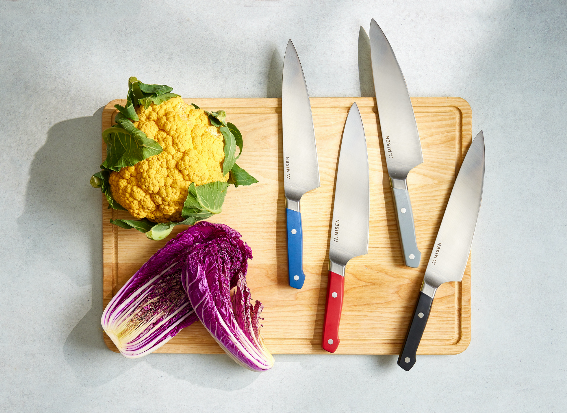 Wood cutting board with five Misen chef knives with colored handles, yellow cauliflower, and halved purple cabbage.