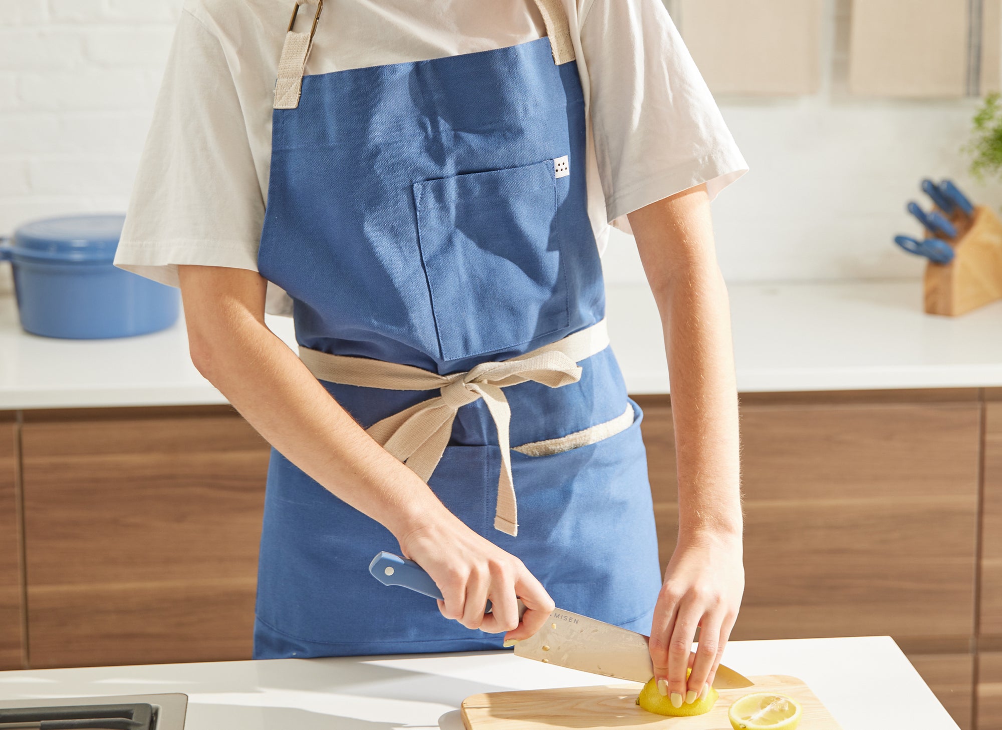 A headless chef wearing a blue Misen apron uses a blue Misen Chef’s Knife to slice lemons on a wood cutting board in a kitchen.