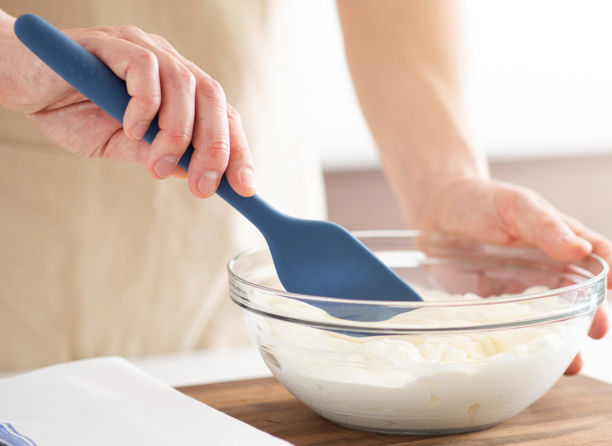 {{blue,black,gray}} Hand using a Blue Misen Mixing Spatula to stir dough in a glass bowl on a kitchen countertop.