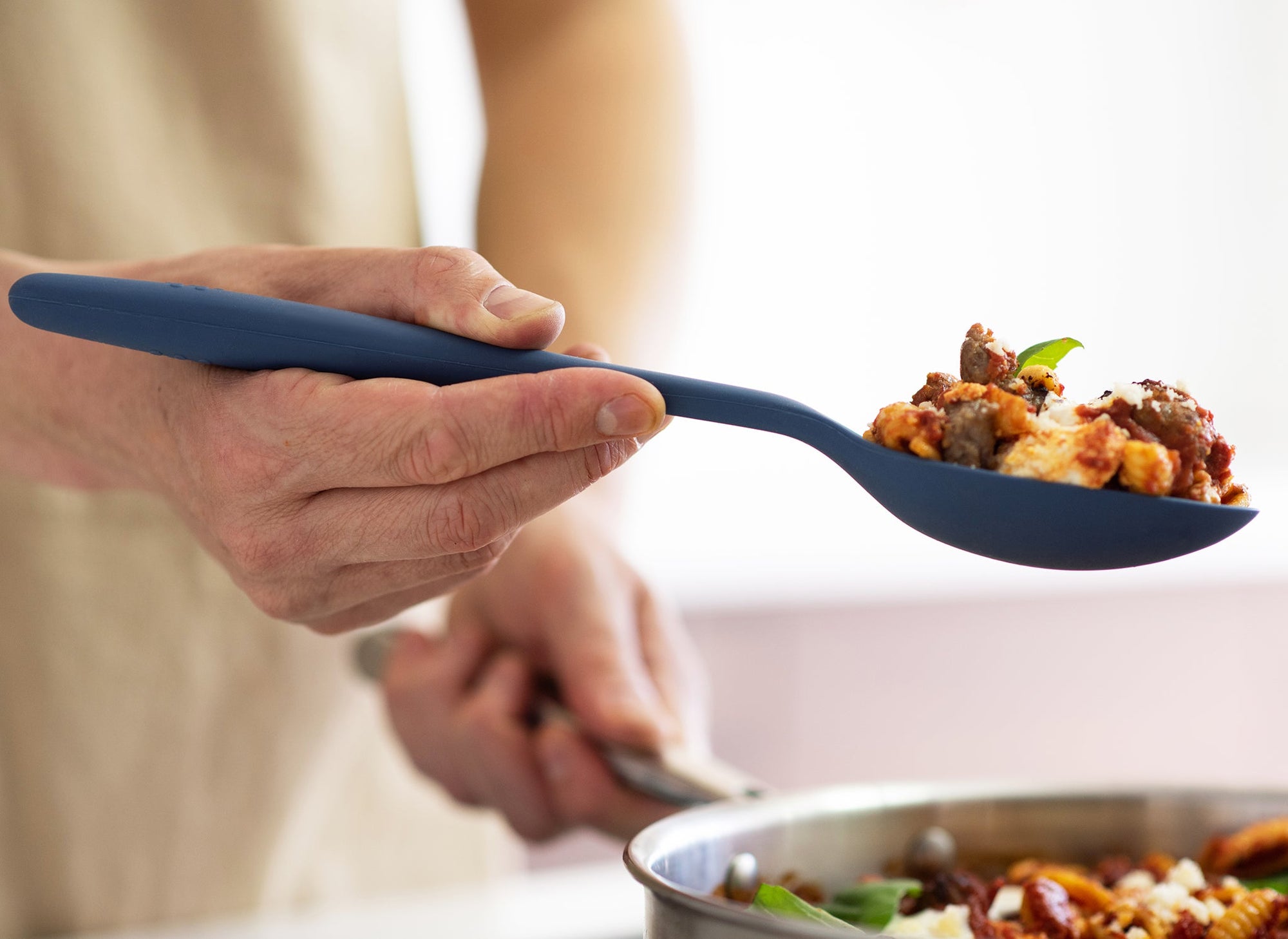 {{blue,black,gray}} A hand using a Blue Misen Mixing Spoon to lift a spoonful of pasta with sausage, basil, and red sauce from a steel pan on a stovetop.