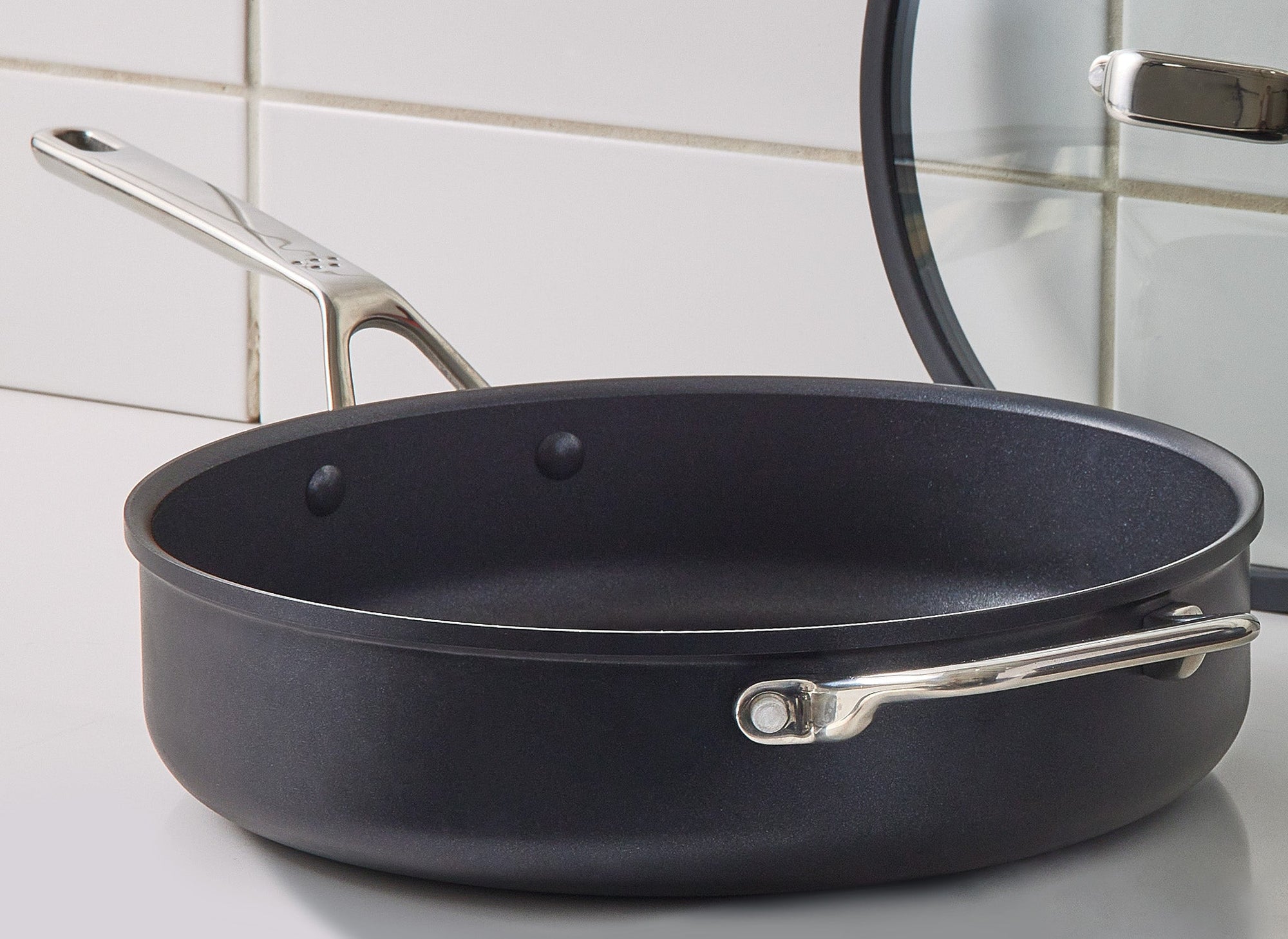 A side-angle view of the Misen 3 QT Nonstick Sauté Pan on a white kitchen counter. The pan’s lid rests against the wall at the edge of the counter.