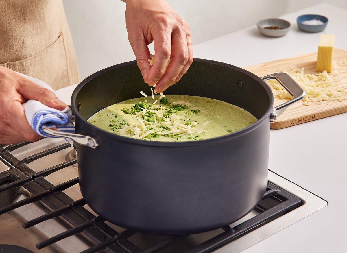 A chef sprinkles cheese in a broccoli soup inside a Misen 8 QT Nonstick Stockpot, which sits on a stovetop.