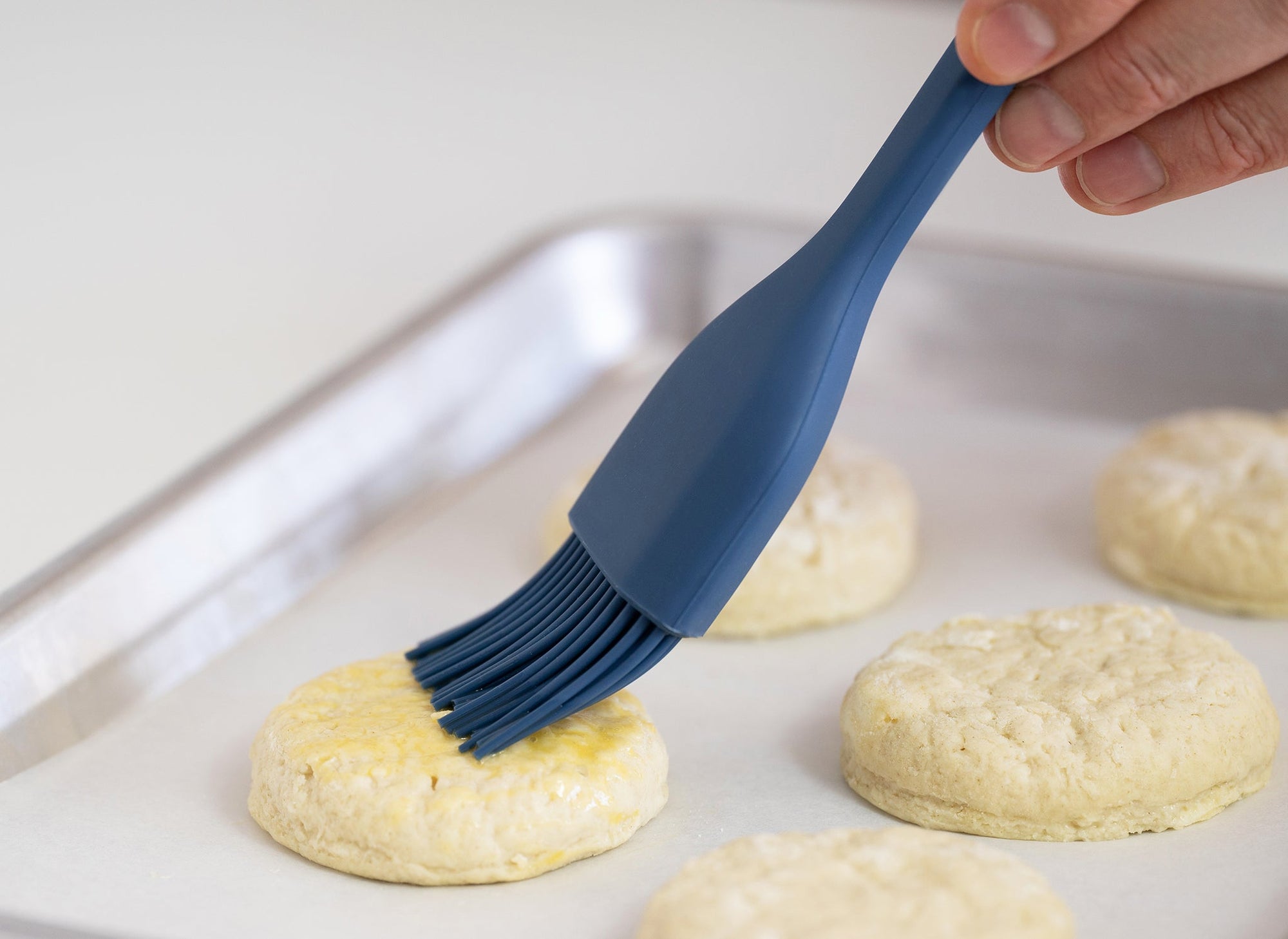 {{blue,black,gray}} Blue Misen Pastry Brush spreading melted butter on biscuits in a baking sheet lined with white paper.