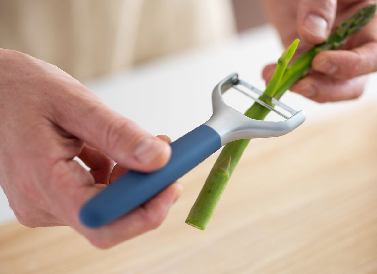 {{blue,black,gray}} A pair of hands using a Blue Misen Peeler to peel the exterior a piece of green asparagus.