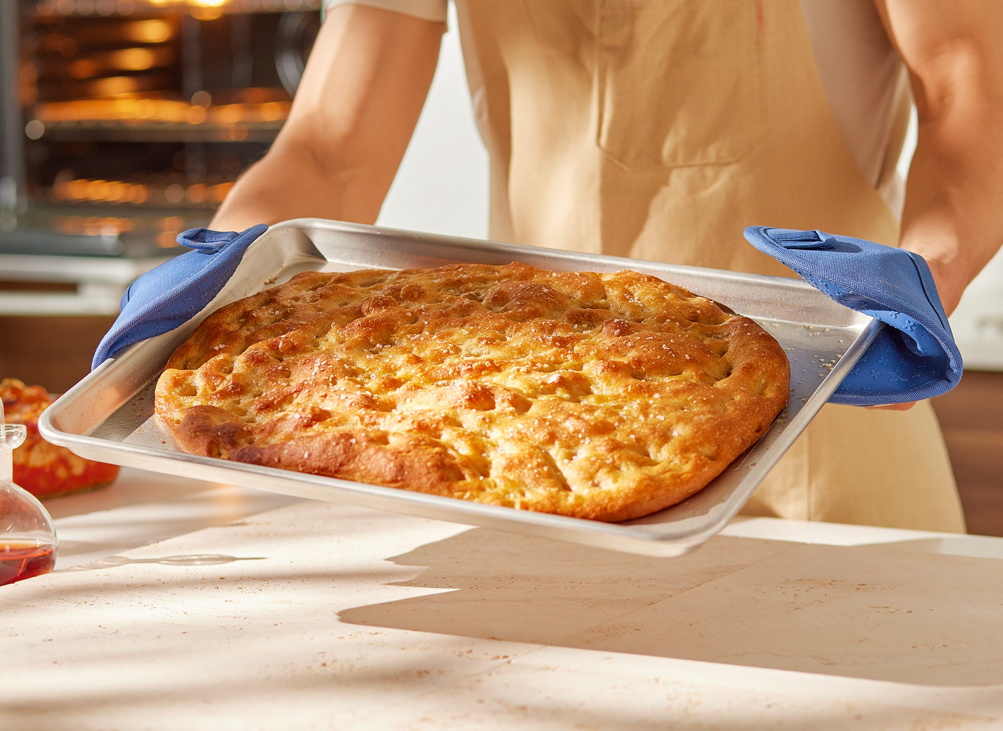 A chef uses two Misen Pot Holders to grip a Misen Sheet Pan filled with a freshly-baked slab of focaccia bread. 