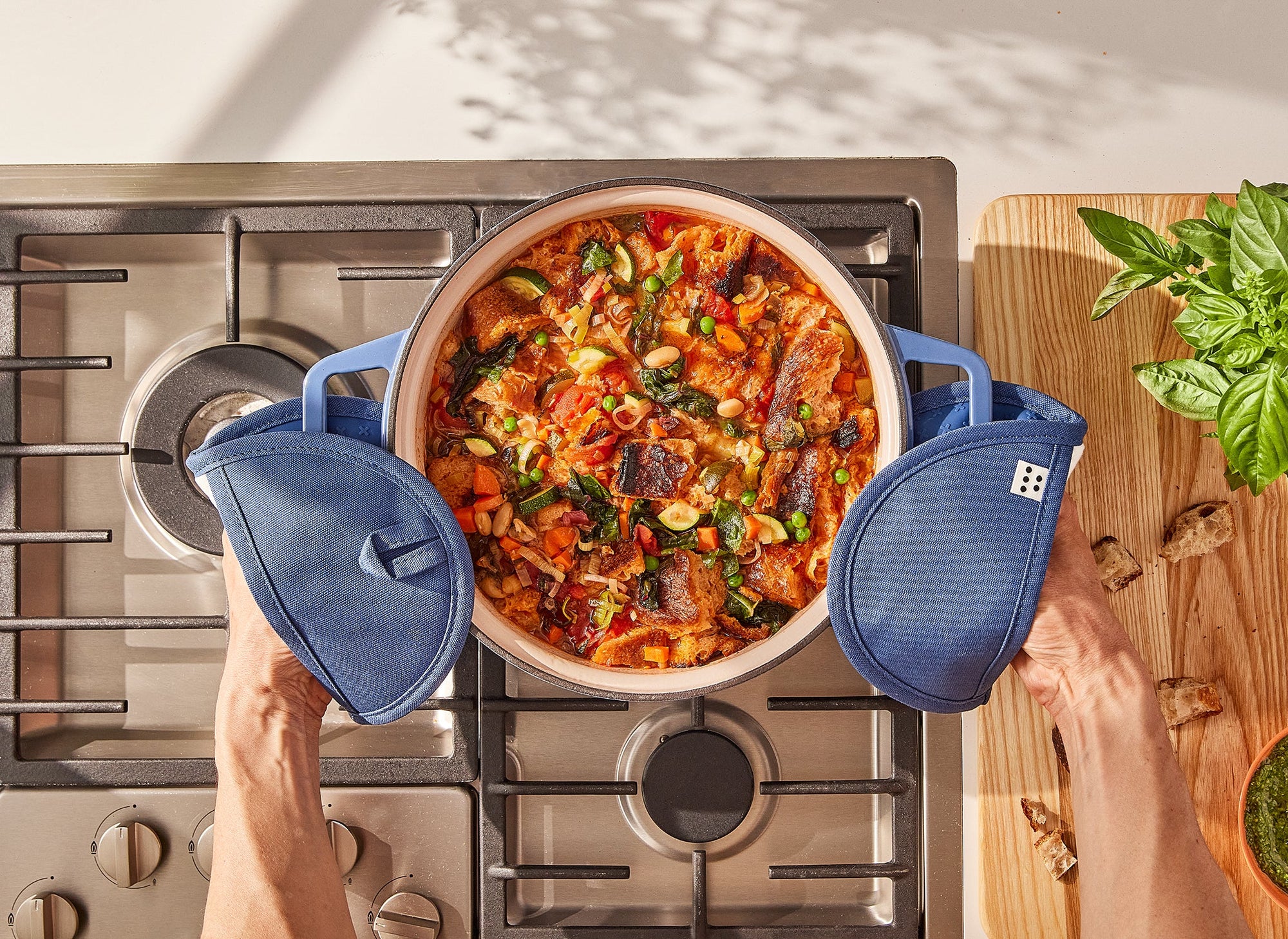 A bird’s eye view of two hands inside Misen Pot Holders grabbing the handles of a blue Misen Dutch Oven, which sits on a stovetop filled with bean and vegetable soup. 