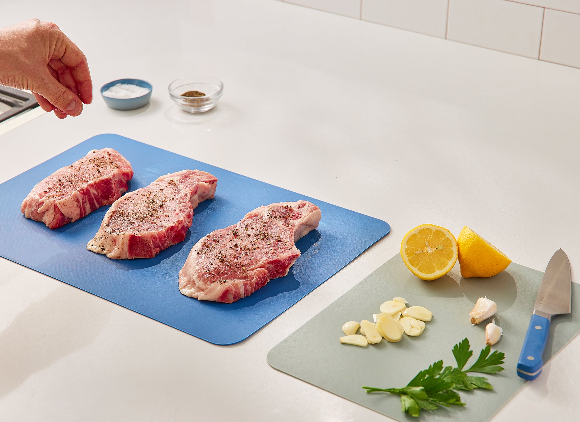 {{plastic}} A hand seasons three raw steaks on a large Misen Cutting Sheet, while sliced lemon, garlic, and parsley rest nearby on a small Misen Cutting Sheet.