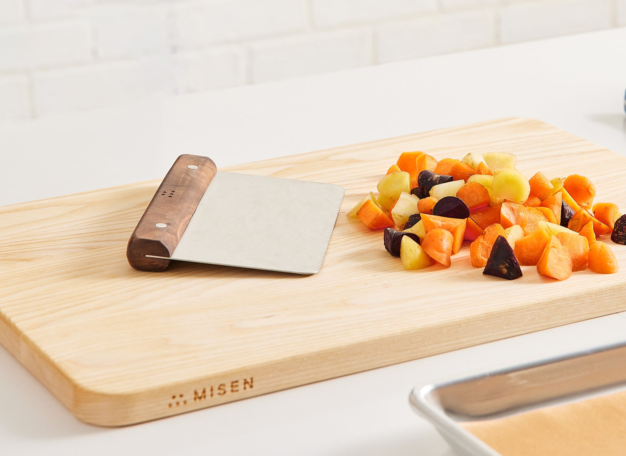 {{walnut,blue}} A Misen Bench Scraper with walnut handle rests atop a Misen Wood Cutting Board next to chopped root vegetables.