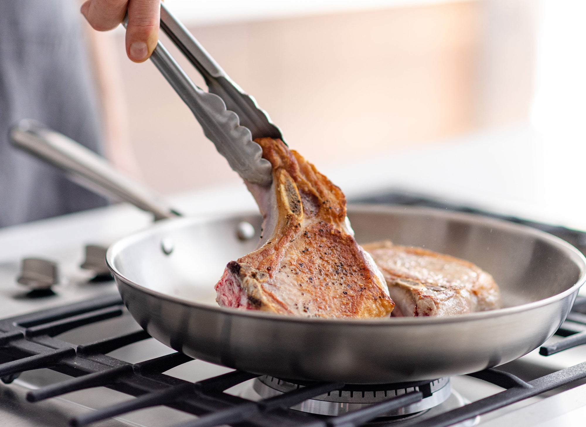 A pair of hands uses tongs to flip a seared bone-in pork chop in a Misen Stainless Steel Pan on a stovetop.